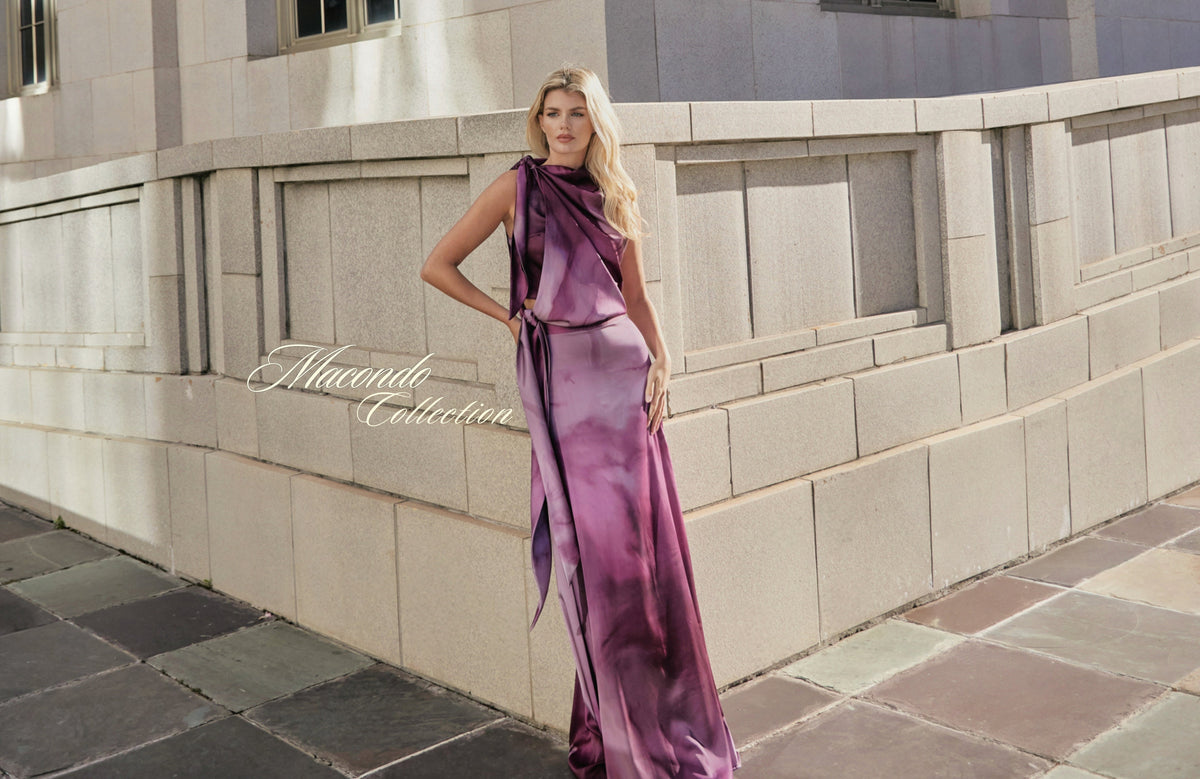 Tinta Latina Macondo campaign: a model wearing a purple draped top and flowing skirt beside a stone building